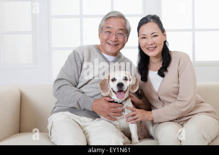 Couple assis sur le canapé avec un chien et sourit joyeusement à l'appareil photo, Banque D'Images