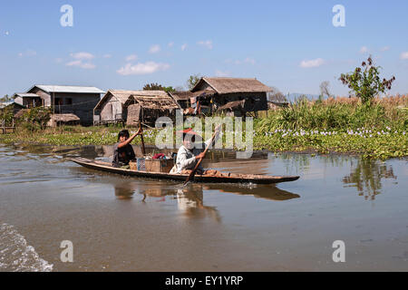 Des maisons sur pilotis traditionnelles au Lac Inle, à l'avant deux femmes locales dans un bateau en bois, de l'État de Shan, Myanmar Banque D'Images