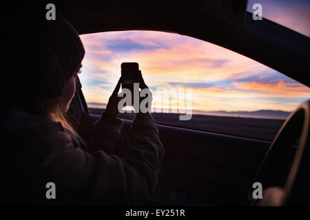 Young woman taking photograph of coucher du soleil avec le smartphone de l'intérieur de voiture Banque D'Images