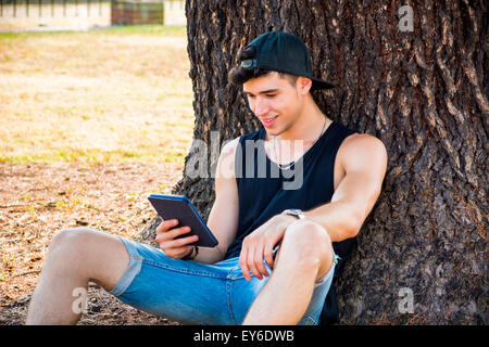 Jeune homme séduisant, lecture sur Ebook Reader ou Holding a Tablet Computer, relaxant sur terrain herbeux au parc, se penchant aga Banque D'Images