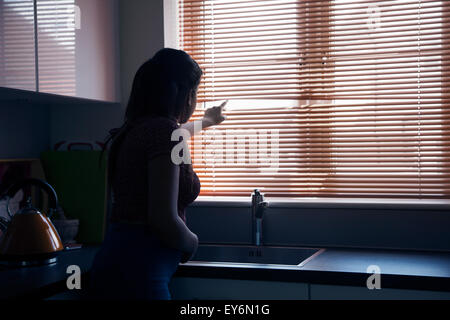 Jeune femme regardant à travers une fenêtre aveugle, silhouette, vue arrière. Banque D'Images