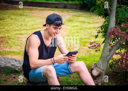 Jeune homme séduisant, lecture sur Ebook Reader ou Holding a Tablet Computer, relaxant sur terrain herbeux au parc, se penchant aga Banque D'Images