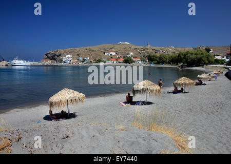 Rangée de parasols en chaume et des nageurs à la plage de sable d''Agios Efstratios ayant le village pittoresque en arrière-plan. Banque D'Images