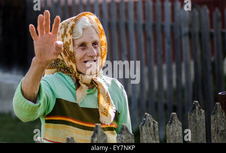 Vieille femme campagne roumaine femme âgée vague joyeusement de sa cour dans un cadre rural, vêtue d'une tenue traditionnelle pendant une journée ensoleillée Banque D'Images