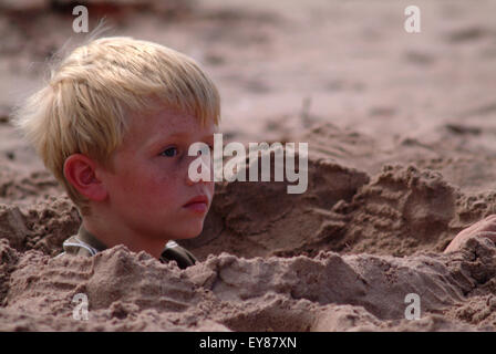 Garçon blond looking pensive, assis dans un trou de sable profond Banque D'Images