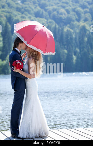 Couple marié debout sur une passerelle, au bord du lac, s'embrasser sous un parapluie rose. Banque D'Images