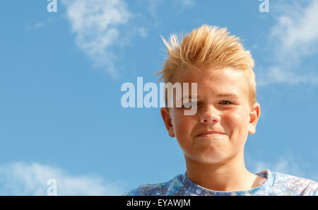 Blonde teenage boy (13 ans) avec un sourire cheveux hérissés ou sourire à la cool contre un ciel bleu avec des nuages blancs Banque D'Images