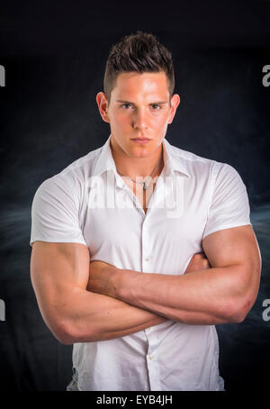 Beau jeune homme élégant avec chemise, looking at camera, isolé sur fond noir, avec l'expression sévère et les bras croisés sur la poitrine Banque D'Images