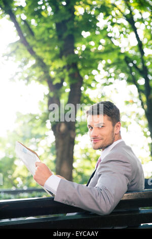 Smiling businessman assis sur le banc avec du papier journal et à la recherche à l'extérieur de l'appareil photo Banque D'Images