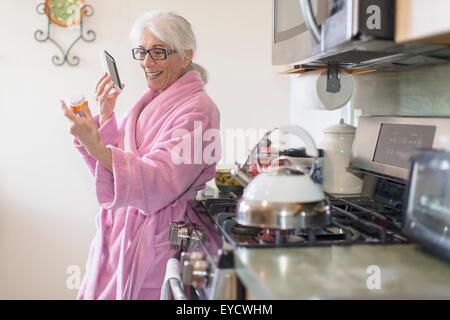Senior woman standing in kitchen holding médicaments bouteille tout en parlant sur le smartphone. Banque D'Images