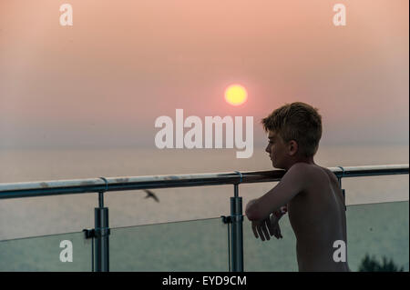 Un garçon regarde le coucher de soleil depuis un mur de verre en vacances Banque D'Images