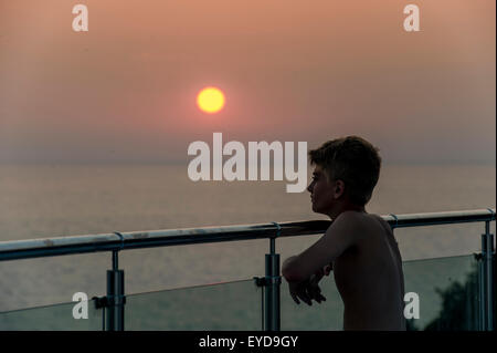 Un garçon regarde le coucher de soleil depuis un mur de verre en vacances Banque D'Images