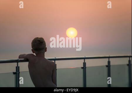 Un garçon regarde le coucher de soleil depuis un mur de verre en vacances Banque D'Images