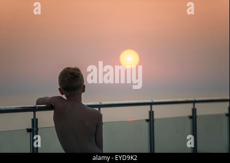 Un garçon regarde le coucher de soleil depuis un mur de verre en vacances Banque D'Images