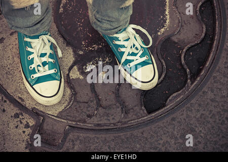 Adolescent en baskets. Pieds de gumshoes rusty se tiennent sur un couvercle de trou d'urbains. Photo Gros Plan avec focus sélectif et vintage Banque D'Images