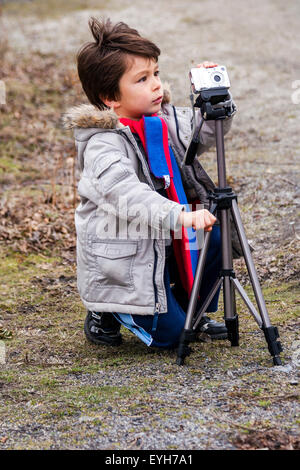 Garçon à l'aide d'une caméra montée sur un trépied à l'extérieur sur la bonne voie. Caucasien enfant, 6-7 ans, les cheveux bruns, porte manteau et écharpe. L'hiver, campagne. Banque D'Images