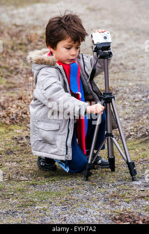 Garçon à l'aide d'une caméra montée sur un trépied à l'extérieur sur la bonne voie. Caucasien enfant, 6-7 ans, les cheveux bruns, porte manteau et écharpe. L'hiver, campagne. Banque D'Images