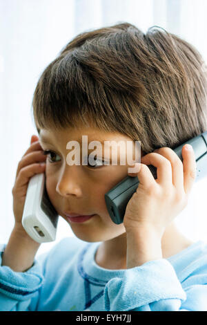 Garçon, 6-7 ans, la tenue de deux téléphones, un pour chaque oreille tandis qu'il écoute. Close up, la tête et les épaules. Caucasien enfant, les cheveux bruns. À l'intérieur. Banque D'Images