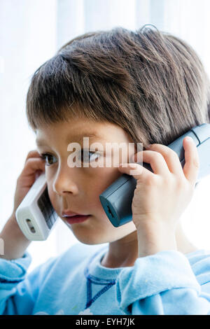 Garçon, 6-7 ans, la tenue de deux téléphones, un pour chaque oreille tandis qu'il écoute. Close up, la tête et les épaules. Caucasien enfant, les cheveux bruns. À l'intérieur. Banque D'Images