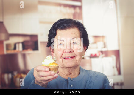 Hispaniques âgées happy woman wearing blue sweater assis en face d'une caméra montrant avec cupcake topping crème Banque D'Images