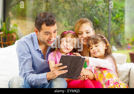 Portrait de famille du père, mère et deux filles assis ensemble dans canapé jouant joyeusement avec tablet Banque D'Images