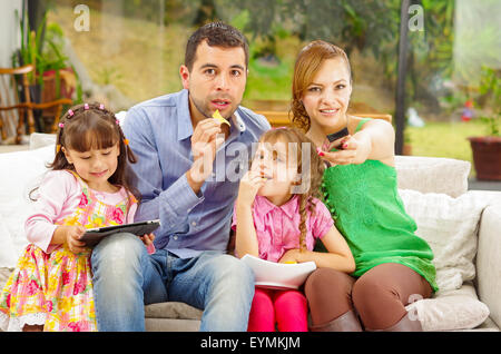 Portrait de famille du père, mère et deux filles assis ensemble dans le canapé à jouer avec comprimé tout en profitant des nachos heureusement souriant pour appareil photo. Banque D'Images