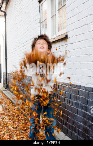 Garçon caucasien enfant, s'amuser dans un mur par la route, en prenant des poignées d'automne les feuilles tombées et les jeter en l'air toutes sur lui-même. Banque D'Images