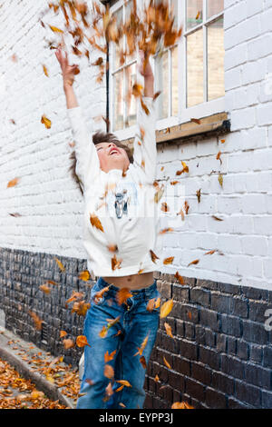 Garçon caucasien enfant, s'amuser dans un mur par la route, en prenant des poignées d'automne les feuilles tombées et les jeter en l'air toutes sur lui-même. Banque D'Images