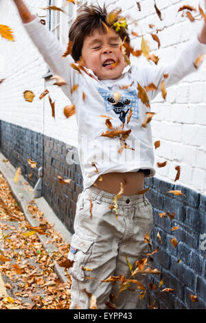 Garçon caucasien enfant, s'amuser dans un mur par la route, en prenant des poignées d'automne les feuilles tombées et les jeter en l'air toutes sur lui-même. Banque D'Images