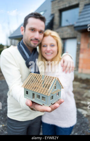 Vue d'un jeune couple heureux de manipuler une miniature house en face de leur future maison Banque D'Images