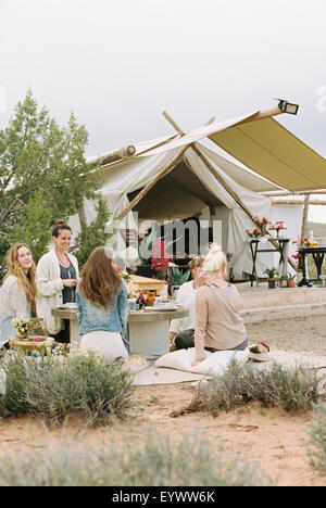 Groupe de femmes amis assis sur le sol autour d'une table dans un désert, une tente dans l'arrière-plan. Banque D'Images
