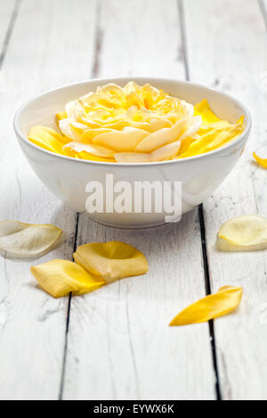 Rose jaune dans un bol d'eau, pétales autour, sur table rustique en bois blanc Banque D'Images