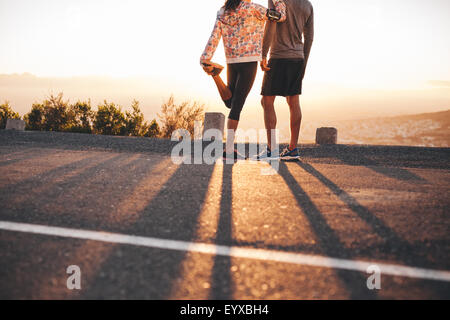 Cropped shot of man and woman standing on hillside road à matin. Femme étendant sa jambe. Couple de coureurs à la recherche à sunri Banque D'Images