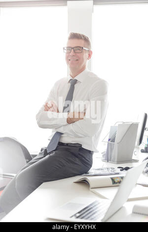 Portrait confident businessman with eyeglasses sitting on desk with arms crossed Banque D'Images