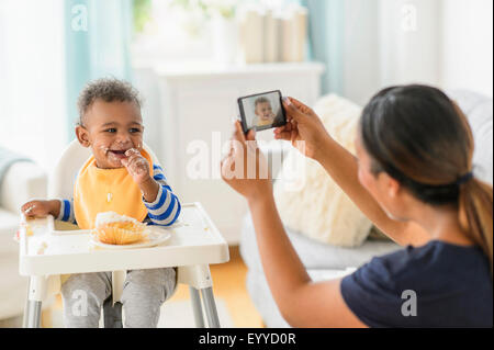 Mixed Race mother photographing messy baby boy in chaise haute Banque D'Images