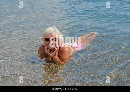 Portrait de senior lady c'est couché dans l'eau peu profonde de la mer de galets de plage. Banque D'Images