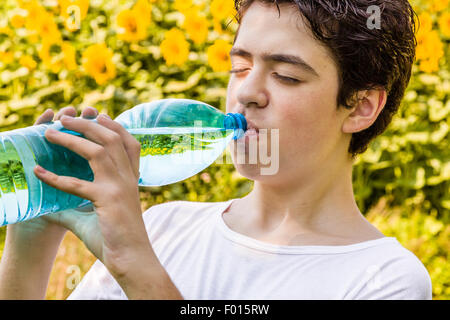 Salon de l'environnement - Caucasian boy dans un champ de tournesols est de boire de l'eau claire d'une bouteille en plastique bleu vert d'étancher sa soif par une chaude journée d'été, lumineux et avec les yeux fermés Banque D'Images