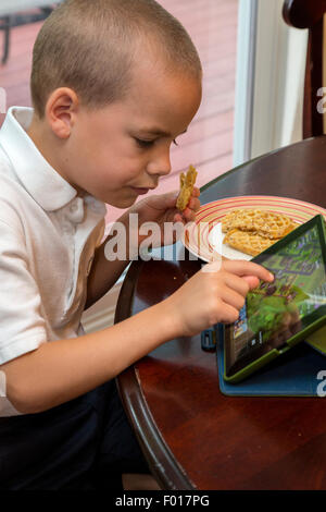 Jeune garçon (7 ans) Joue une partie sur son iPad en mangeant le petit déjeuner. M. Banque D'Images