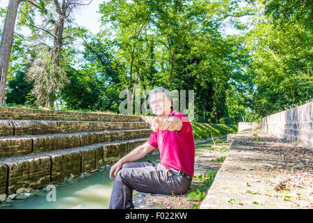 Beau et charismatique Caucasian sportsman de quarante avec cheveux gris polo porter du rouge foncé et un pantalon de lin points tandis que assis sur les marches de l'ancienne cité médiévale du Canal de l'usine en Emilie Romagne en Italie Banque D'Images