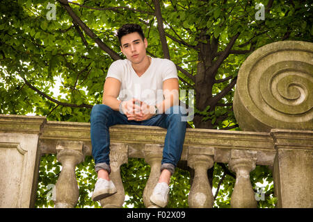 Beau jeune homme assis sur un balcon ou un pont s'appuyant sur la balustrade de pierre souriant à l'appareil photo Banque D'Images