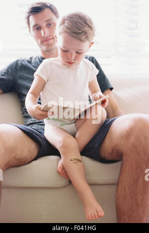 Homme assis sur un canapé avec son fils, tenant un livre, lecture. Banque D'Images