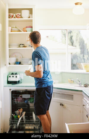 Vue arrière d'un homme debout devant une armoire avec de la vaisselle dans une cuisine. Banque D'Images