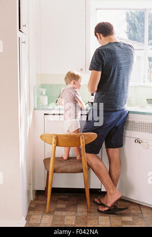 Homme debout dans une cuisine, son fils debout sur une chaise à côté de lui. Banque D'Images