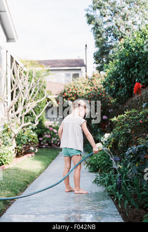 Jeune fille debout sur un chemin dans un jardin, jouant avec un tuyau d'eau. Banque D'Images