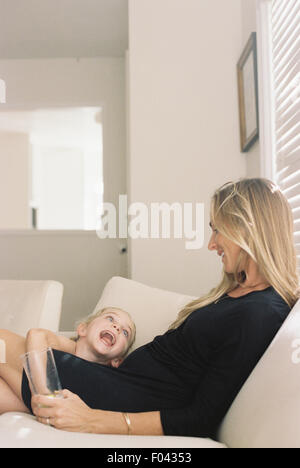 Femme avec de longs cheveux blonds portant une robe noire, assise sur un canapé, tenant un verre, jouant avec sa fille. Banque D'Images