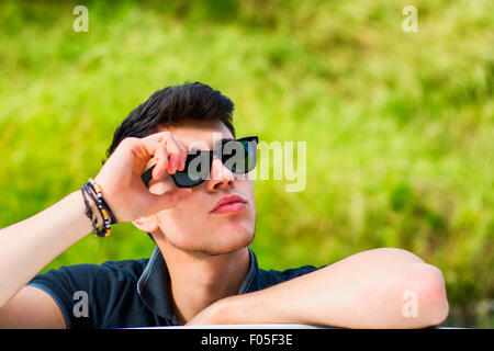 Tourné la tête et les bras de handsome young man looking at camera outdoor, s'appuyant sur une surface plane, tête posée sur la main Banque D'Images