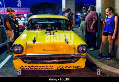 Bellmore, New York, USA. 7 août 2015. Les gens regardent le jaune Chevrolet 1957 muscle car, avec ventilateur (supercharger compresseur à air) qui sort par le trou dans le capot, et rêve américain écrit sous la calandre avant, et stationné sous la voie ferrée surélevée au vendredi soir Car Show s'est tenue à l'Bellmore Long Island Railroad Station Parking. Des centaines de classique, antique, et les voitures personnalisées ont été à l'émission hebdomadaire libre, parrainé par la Chambre de Commerce de l'Bellmores. Banque D'Images