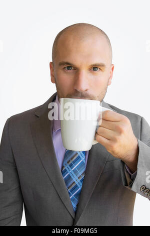 Jeune, beau business man avec le crâne rasé et les cheveux du visage dans un costume gris en sirotant du café blanc tasse et regardant attentivement Banque D'Images