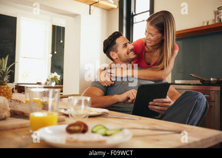 Coup de jeune couple aimant dans la cuisine par table de petit déjeuner le matin. Man using digital table tout en le serrant femme de behin Banque D'Images