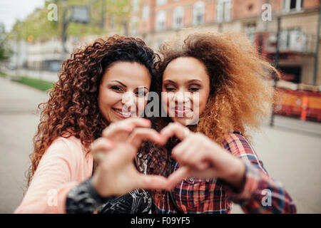 Belles jeunes femmes en forme de coeur avec les doigts. Deux femmes se tenant ensemble à l'extérieur, sur la rue de la ville. Banque D'Images